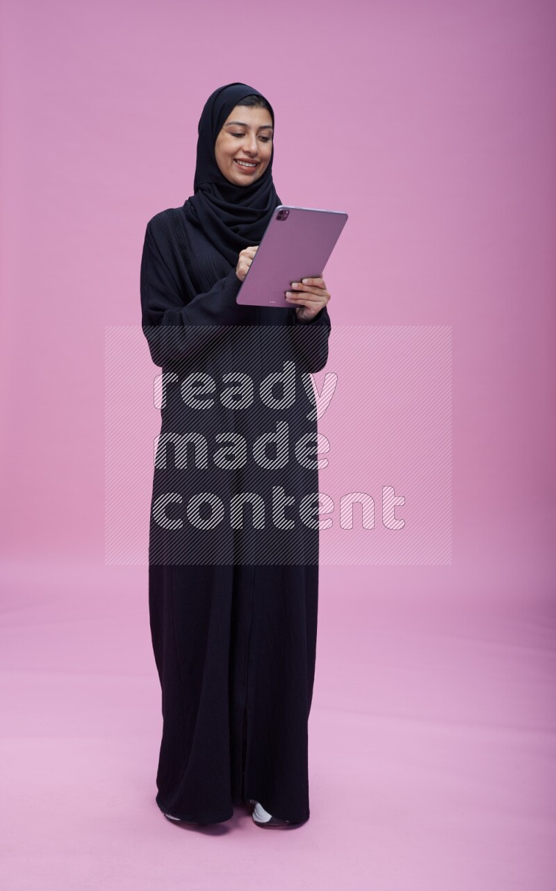 Saudi woman wearing Abaya standing working on tablet on pink background