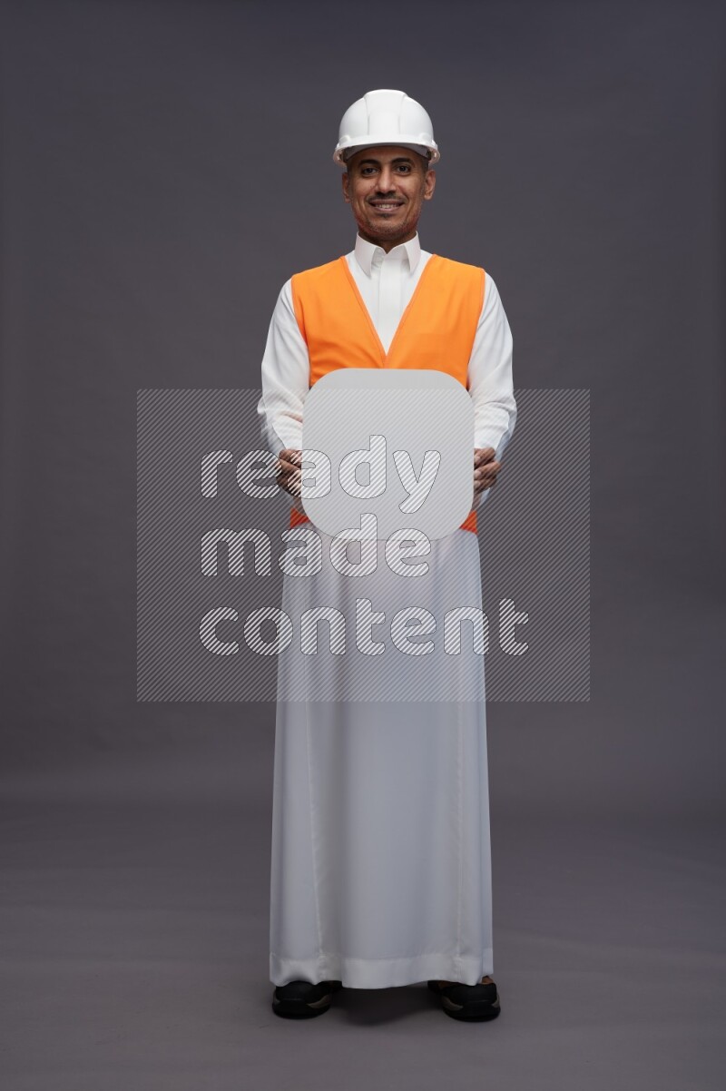 Saudi man wearing thob with engineer vest standing holding social media sign on gray background