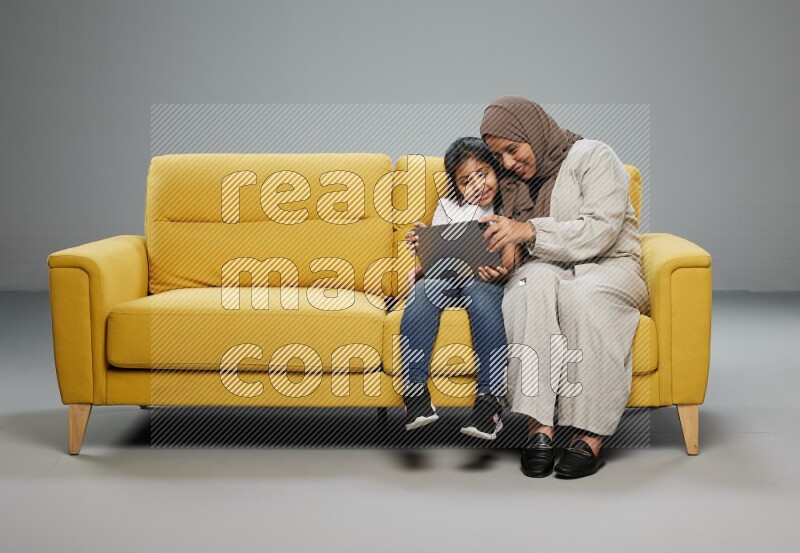 A girl with her mother on yellow sofa and watching on iPad on gray background