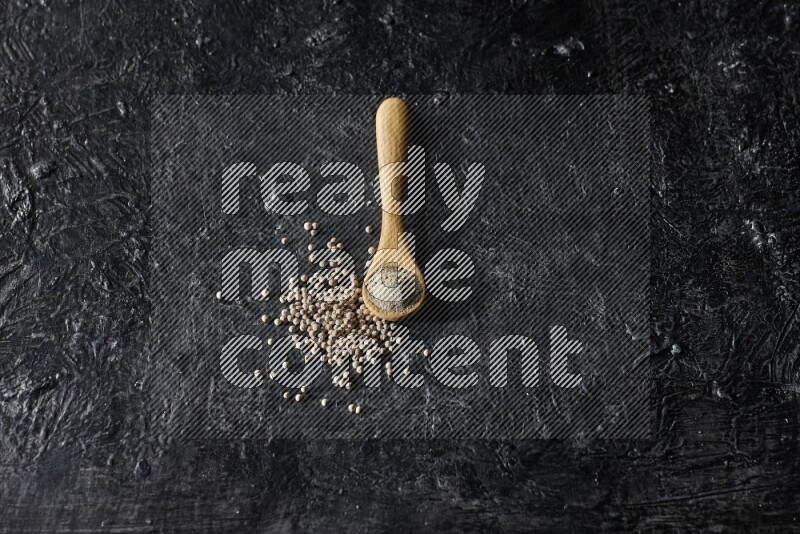 A wooden spoon full of white pepper powder with white pepper beads on textured black flooring