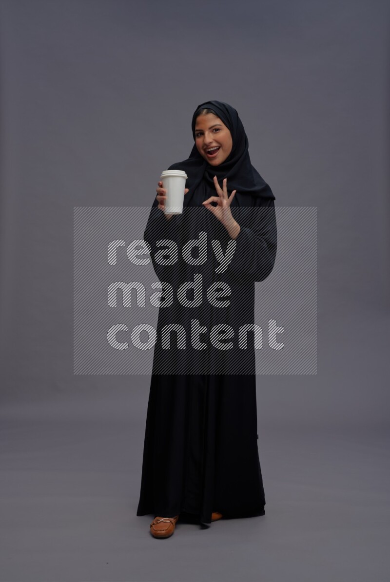 Saudi woman wearing Abaya standing holding paper cup on gray background