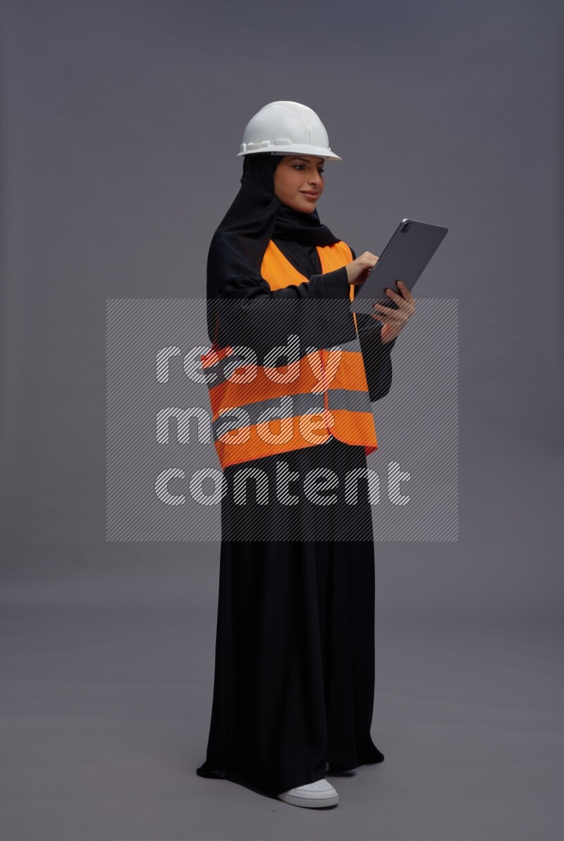 Saudi woman wearing Abaya with engineer vest standing working on tablet on gray background