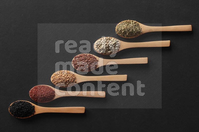 wooden spoons filled with white peppers, mustard seeds, black seeds, garden cress, cumin and flax on black flooring