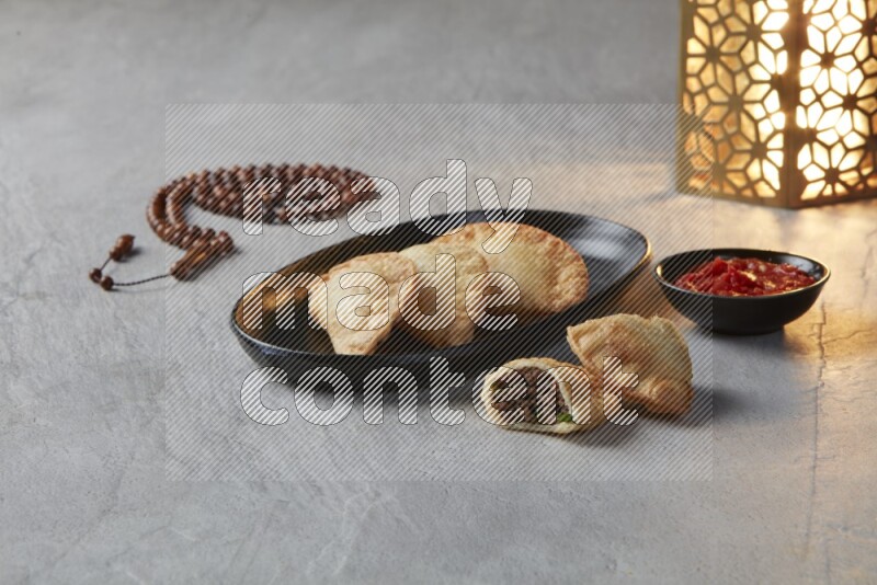 Three fried sambosas in an oval shaped black plate and a red sauce in a black round ramekin with a brown misbaha and a golden lantern on the side on a gray background