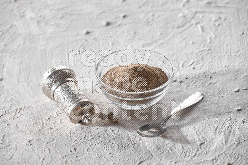 A glass bowl full of black pepper powder and a metal spoon and a turkish metal grinder on textured white flooring