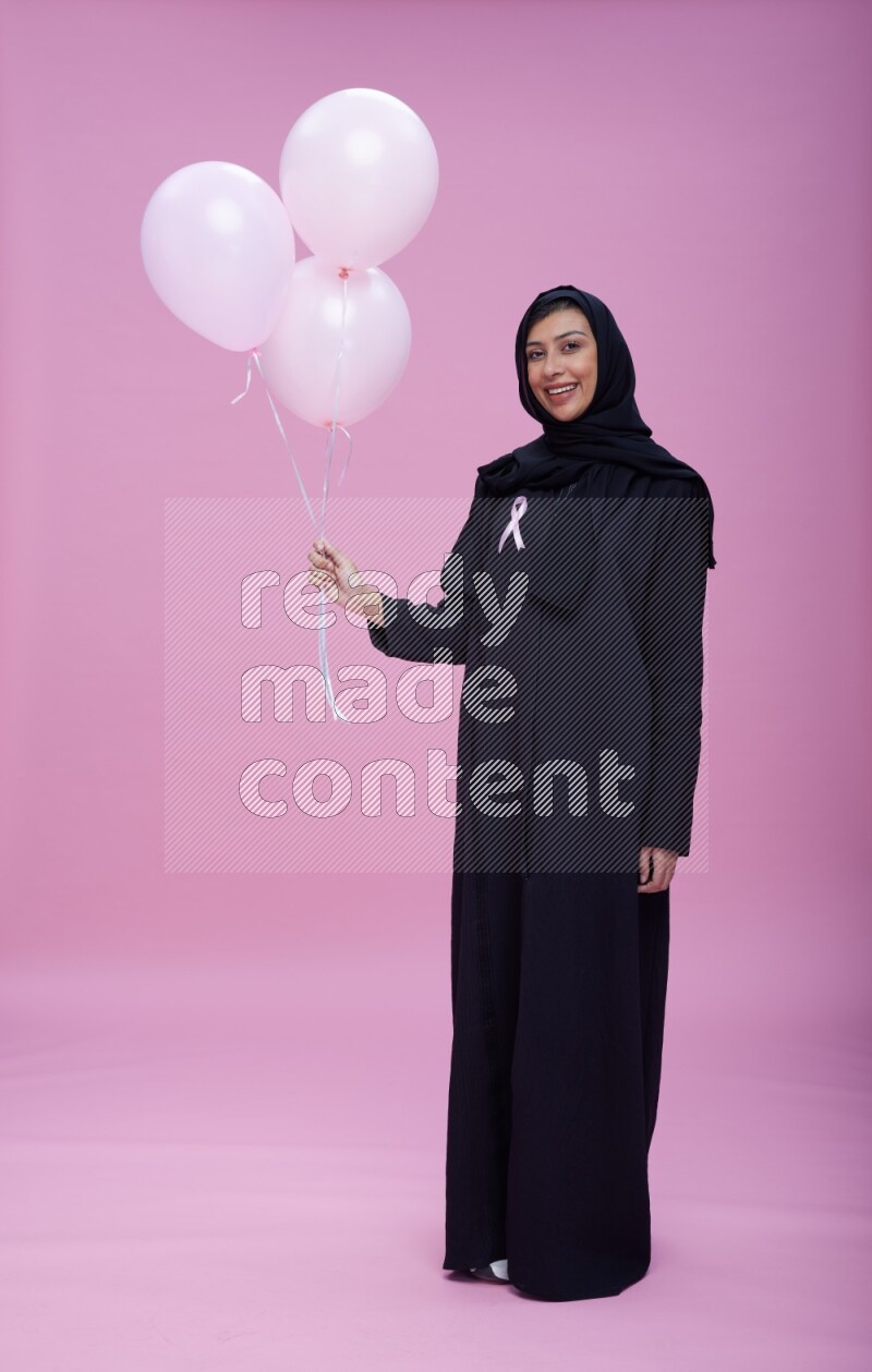 Saudi woman wearing pink ribbon on Abaya standing holding balloons on plain pink background