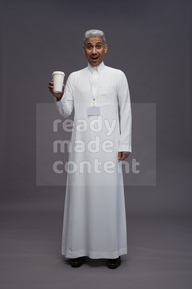 Saudi man wearing thob with neck strap employee badge standing holding paper cup on gray background