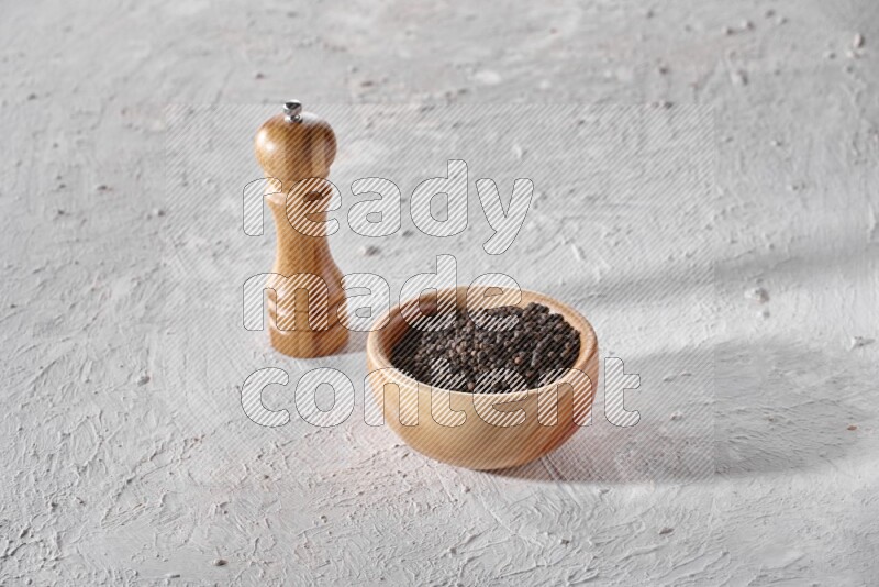 A wooden bowl full of black pepper and a wooden grinder on a textured white flooring