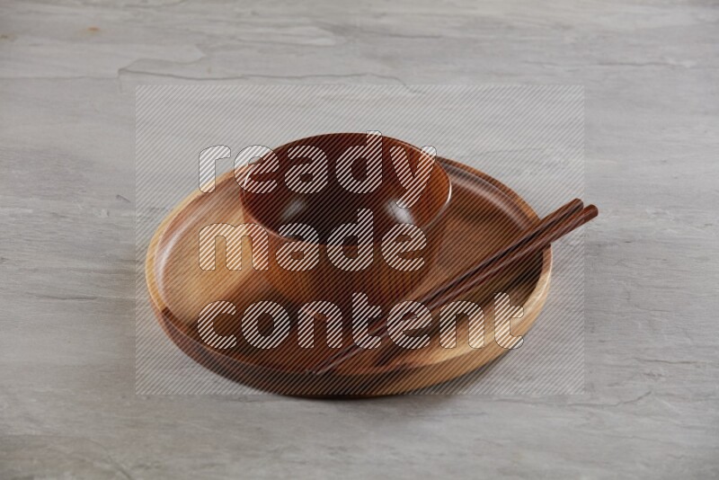 brown wood round bowl on top of brown wood round plate and wood chopsticks, on grey textured countertop