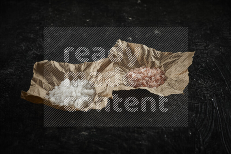 2 crumpled pieces of paper full of pink himalayan salt and white salt on black background