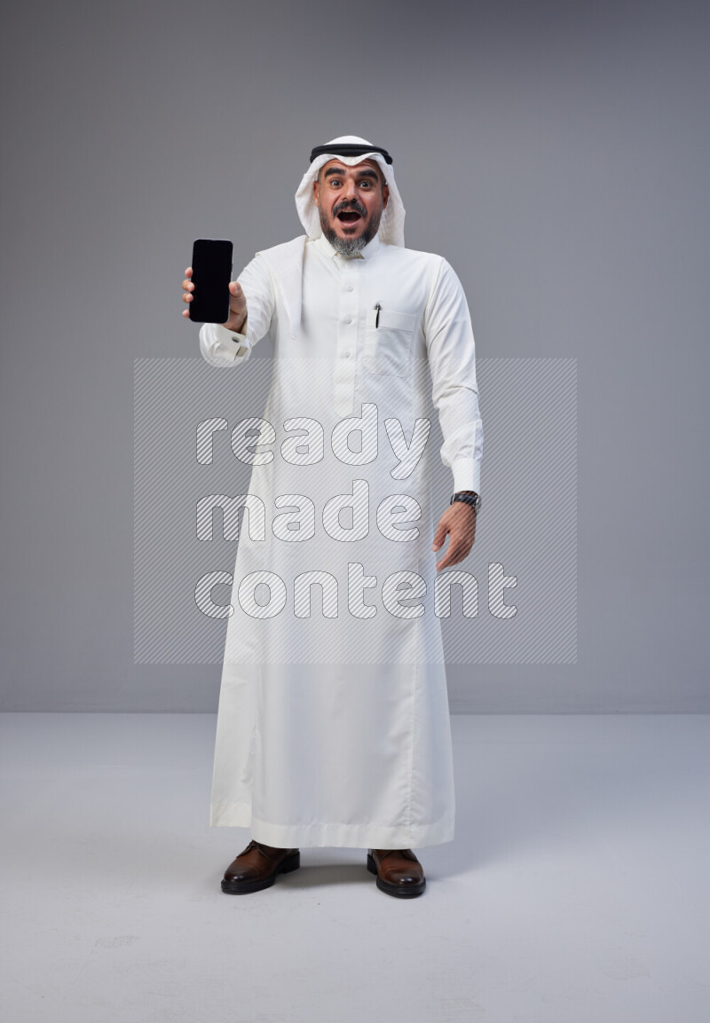 Saudi man Wearing Thob and white Shomag standing showing phone to camera on Gray background