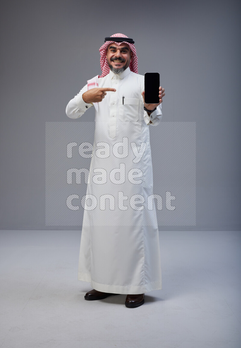 Saudi man Wearing Thob and red Shomag standing showing phone to camera on Gray background