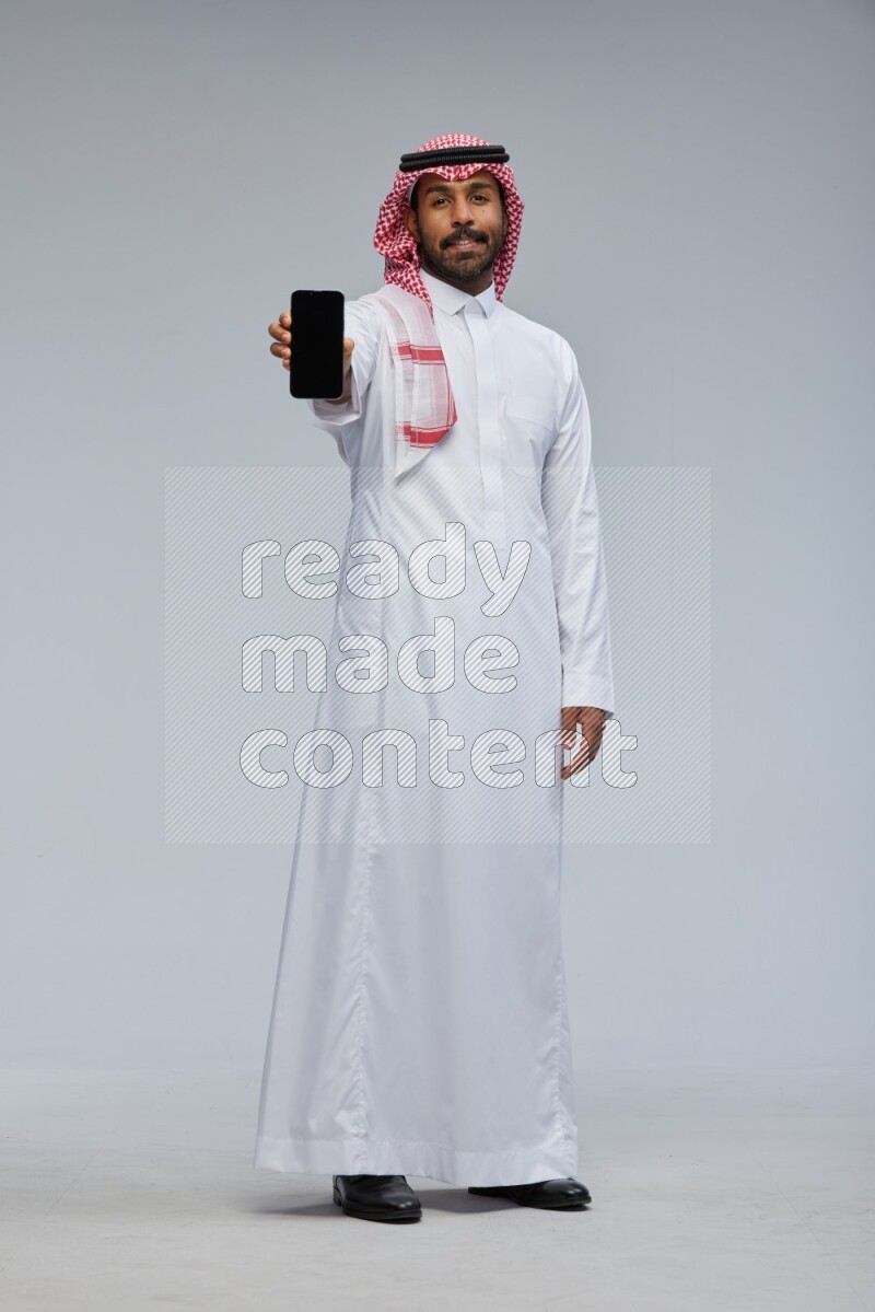 Saudi man Wearing Thob and shomag standing showing phone to camera on Gray background