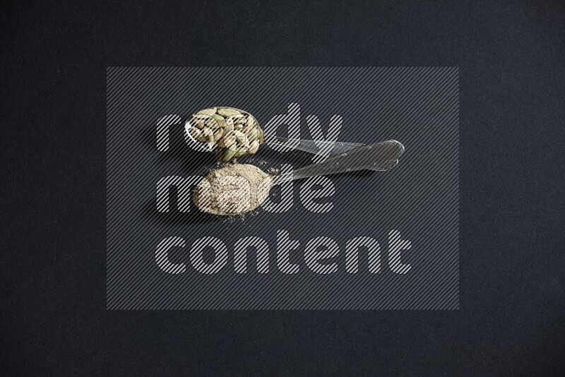 ملعقتان معدنيتان ممتلئتان ببودرة الهيل وحبوب الهيل على خلفية سوداء