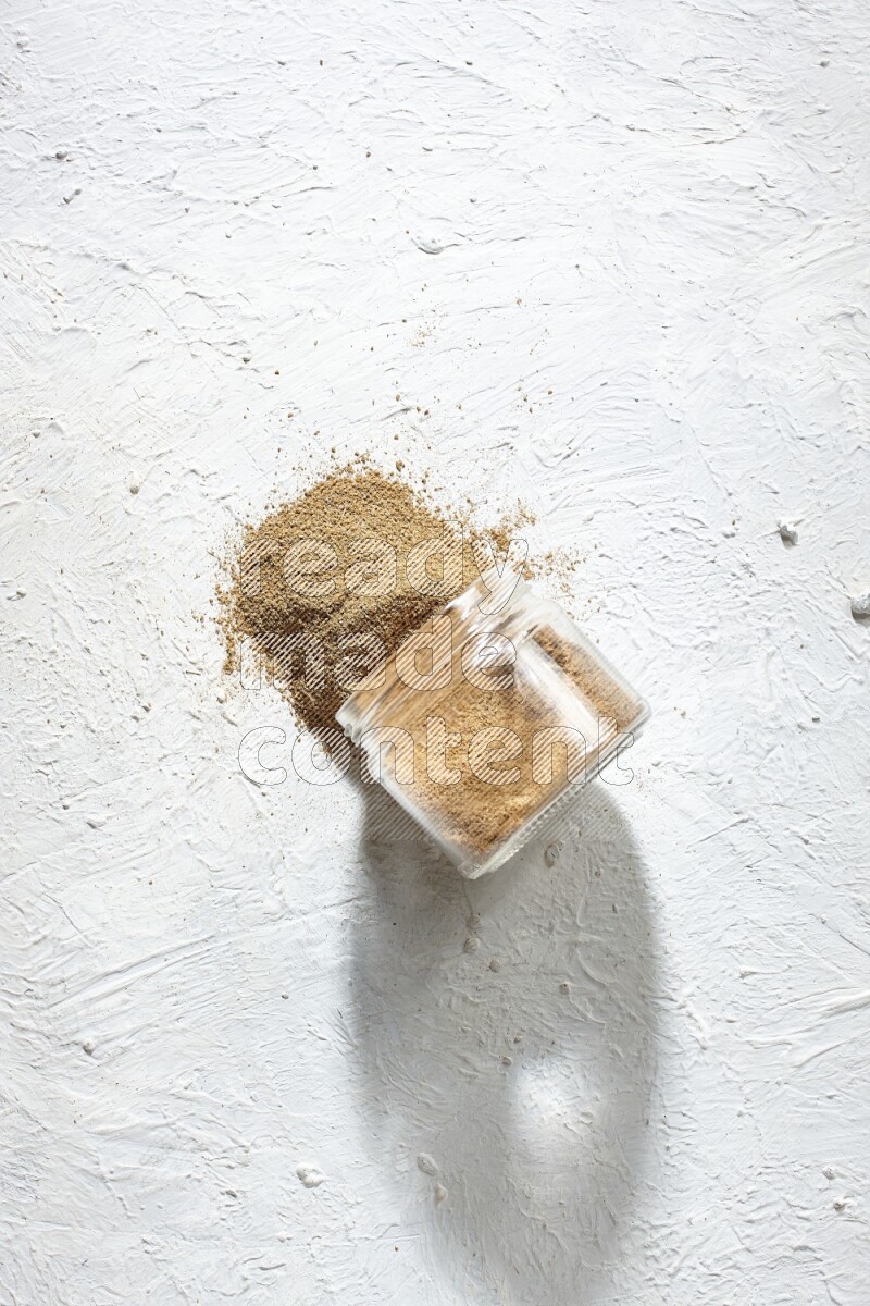 A flipped glass jar full of cumin powder and powder spilled out on textured white flooring