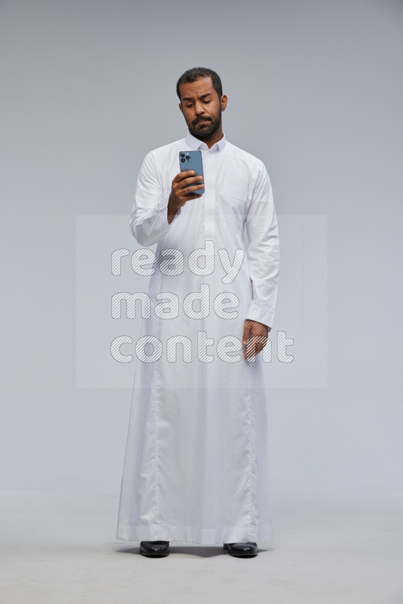 Saudi man wearing Thob standing texting on phone on Gray background