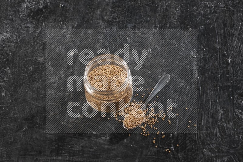 وعاء زجاجي وملعقة معدنية كلاهما ممتلئ ببذور الخردل على أرضية سوداء