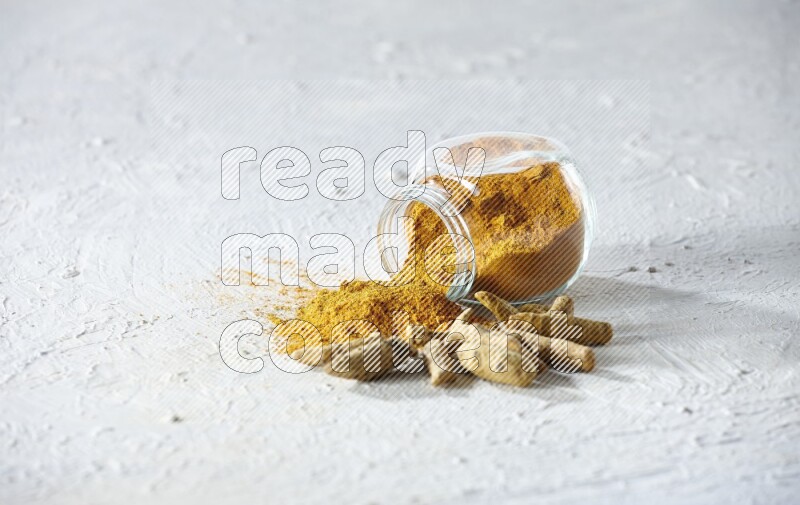 A flipped glass spice jar full of turmeric powder and powder spilled out of it with dried whole fingers on textured white flooring