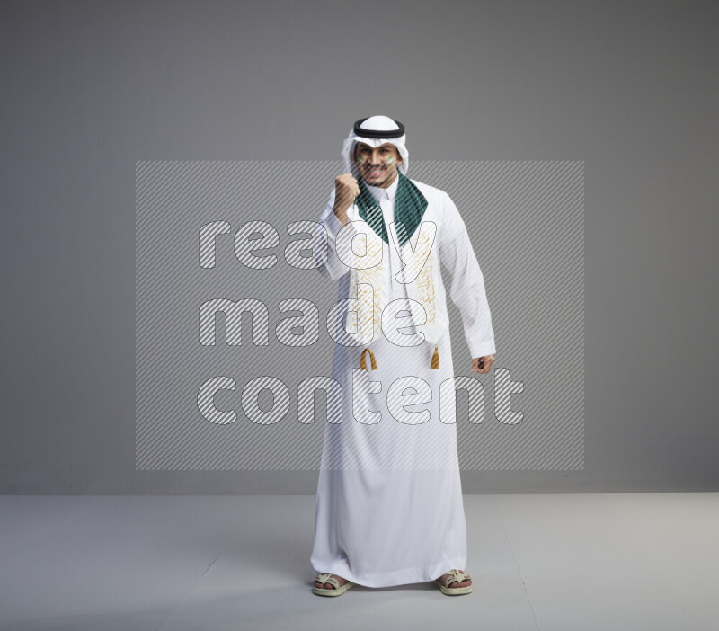 A Saudi man standing wearing thob and white shomag with face painting and Saudi flag scarf on gray background