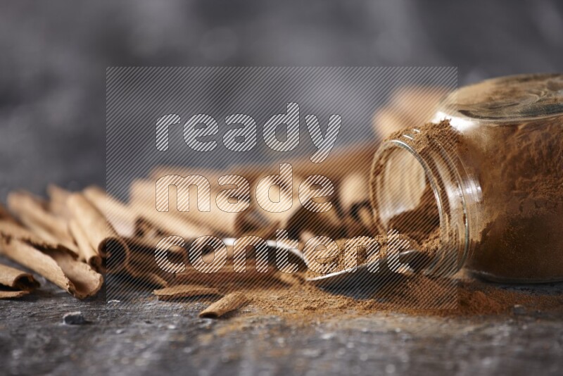 Herbal glass jar full cinnamon powder flipped and a metal spoon full of powder surrounded by cinnamon sticks on textured black background in different angles