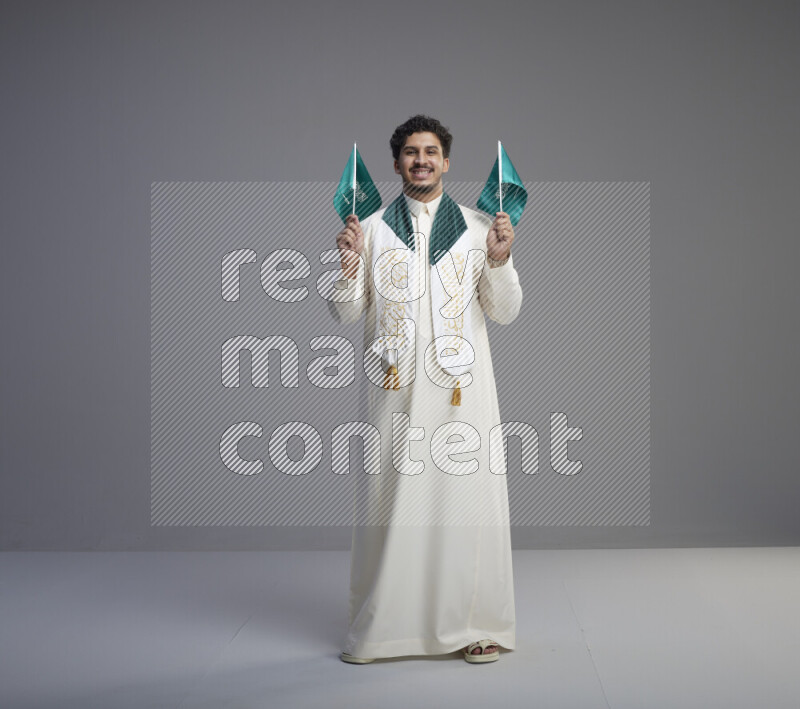 A Saudi man standing wearing thob and saudi flag scarf and holding small saudi flag on gray background
