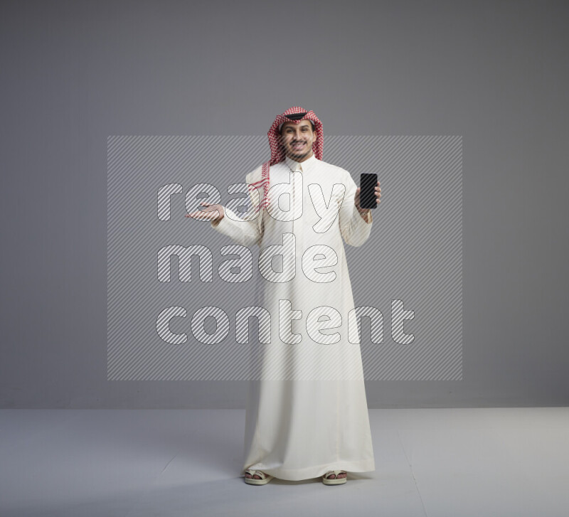 A Saudi man standing wearing thob and red shomag showing phone to camera on gray background
