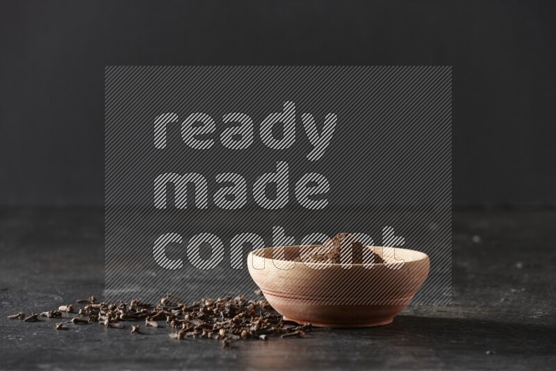 A wooden bowl full of cloves powder on a textured black flooring