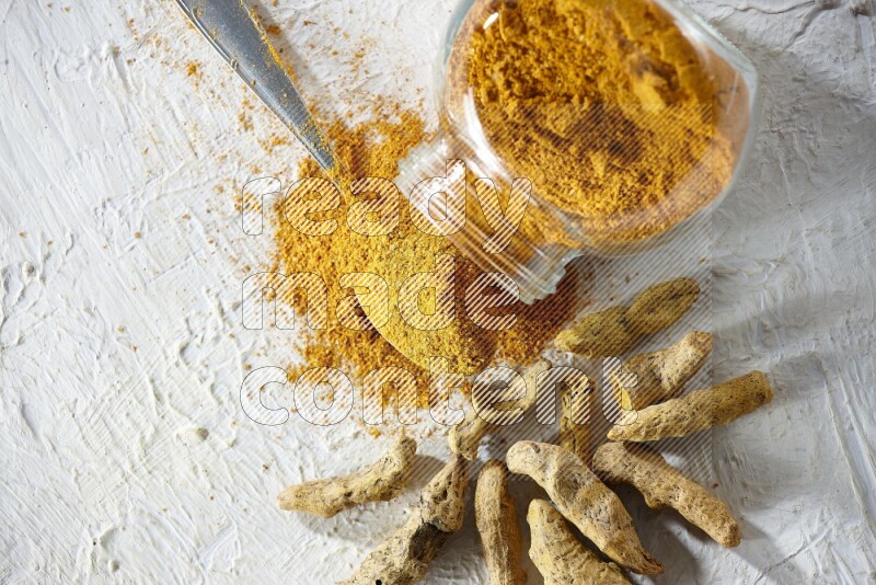 A flipped glass spice jar and metal spoon full of turmeric powder and powder spilled out of it with dried whole fingers on textured white flooring