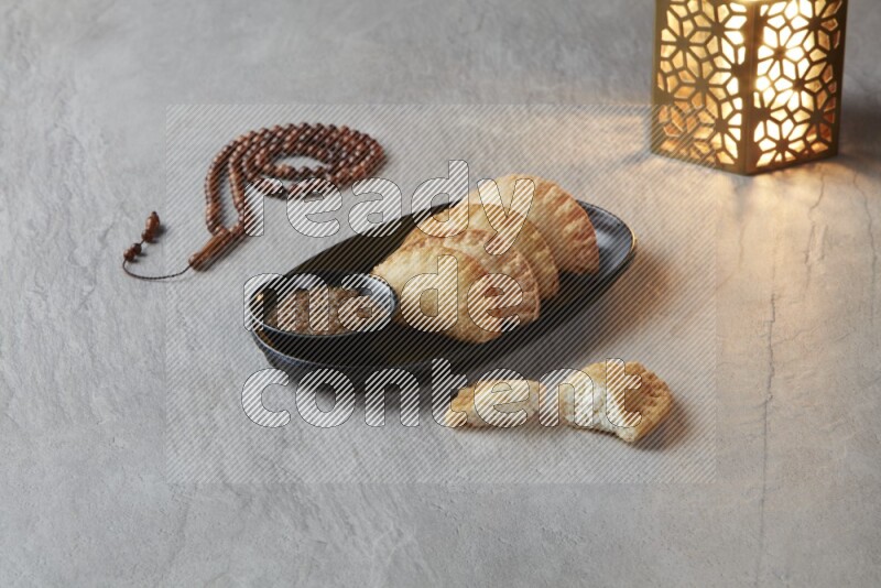 Four fried sambosas in an oval shaped black plate, beside a cut cheese sambosa, a brown misbaha and a golden lantern on a gray background