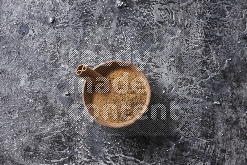 Wooden bowl full of cinnamon powder and a cinnamon stick on a textured black background