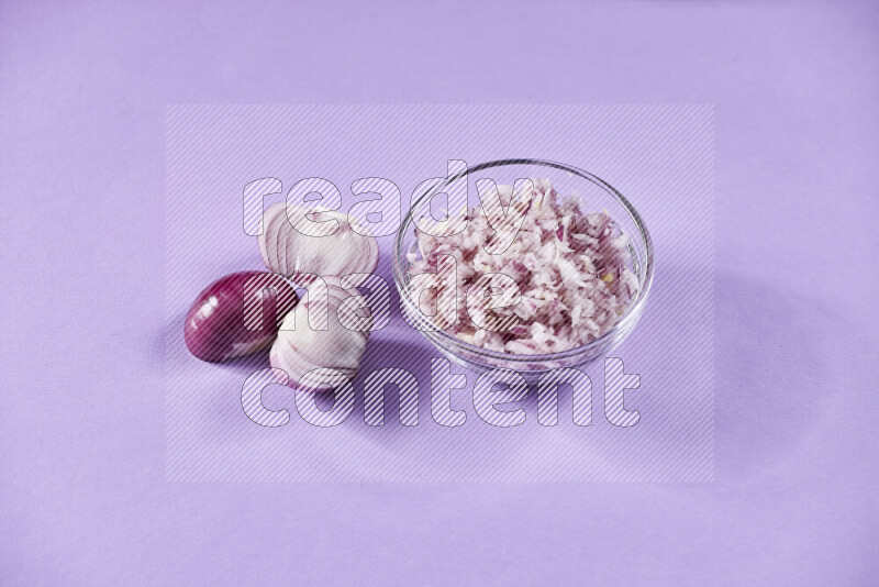 A glass bowl full of chopped red onions with halved onions beside it on purple background