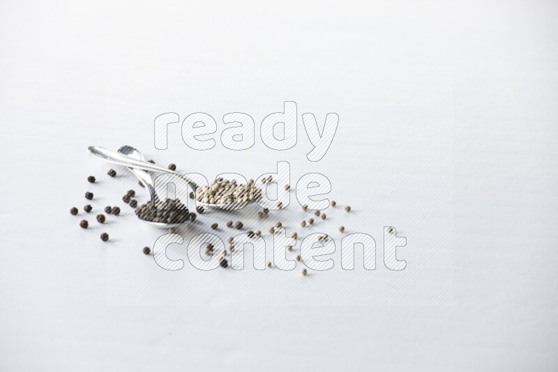 ملعقتان معدنيتان ممتلئتان بحبوب الفلفل الأسود والأبيض مع حبوب متناثرة على أرضية بيضاء