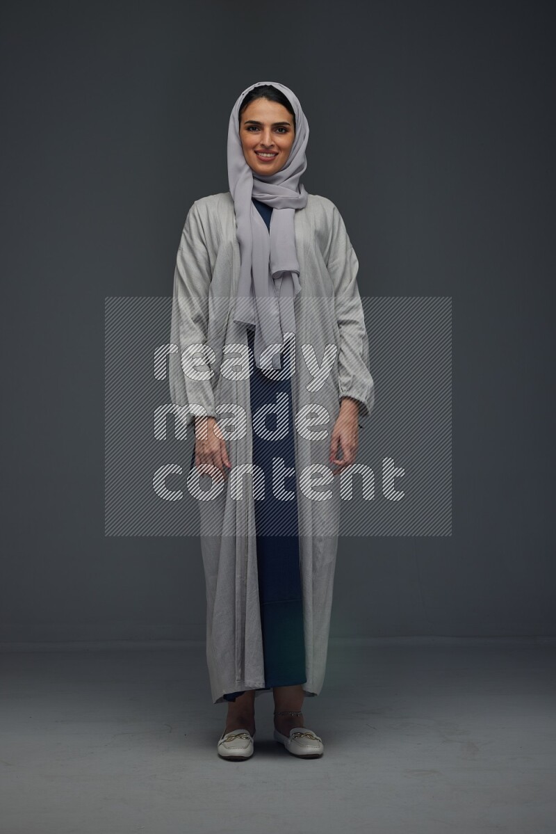 A Saudi woman wearing a light gray Abaya and head scarf standing and making multi poses eye level on a grey background