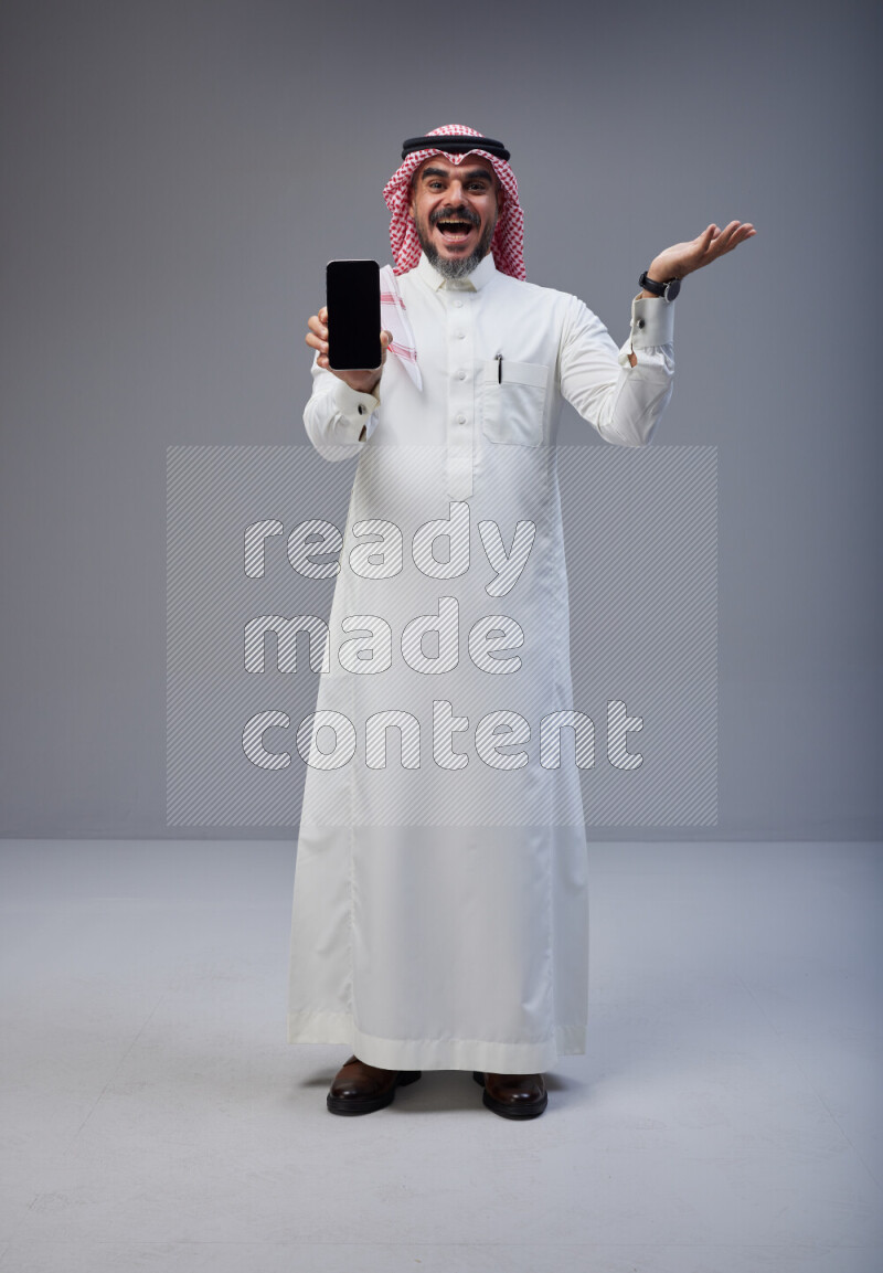Saudi man Wearing Thob and red Shomag standing showing phone to camera on Gray background