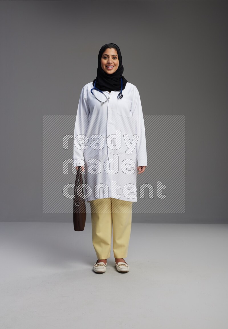 Saudi woman wearing lab coat with stethoscope standing holding bag on Gray background
