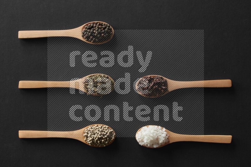 wooden spoons filled with white peppers, cloves, cardamom, salt, black peppers and basil on black flooring