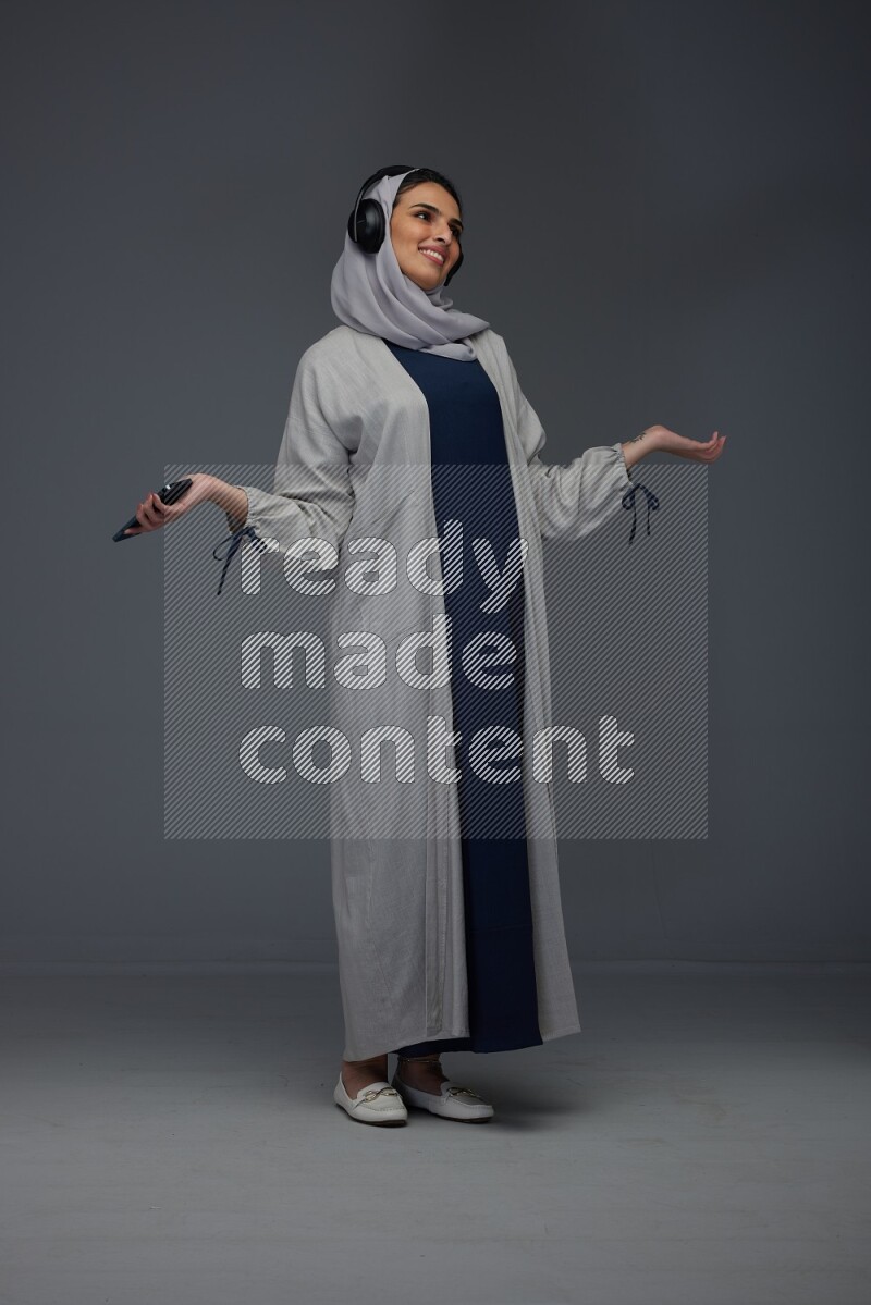 A Saudi woman wearing a light gray Abaya and head scarf standing and listening to music on a grey background