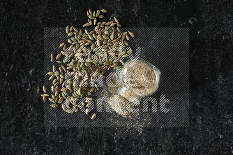 A flipped glass spice jar and a metal spoon full of cardamom powder and cardamom seeds spreaded on textured black flooring