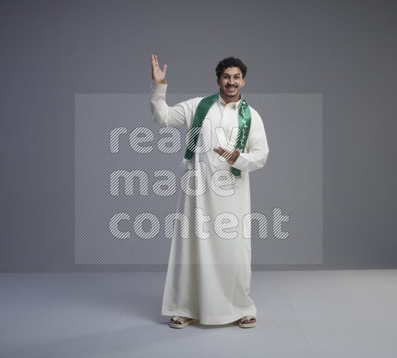 A Saudi man standing wearing thob and Saudi flag scarf on gray background