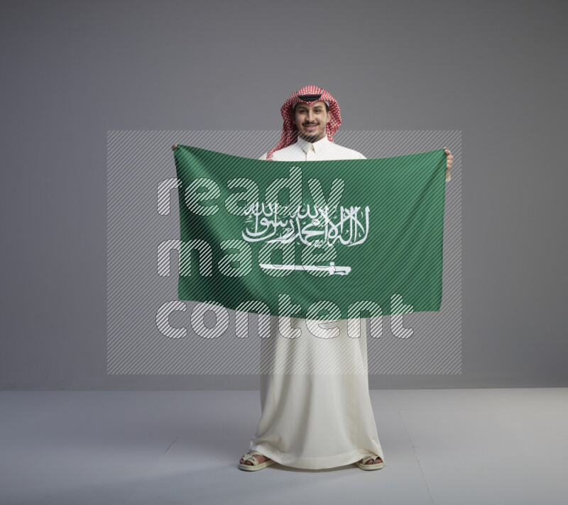 A saudi man standing wearing thob and red shomag holding big saudi flag on gray background