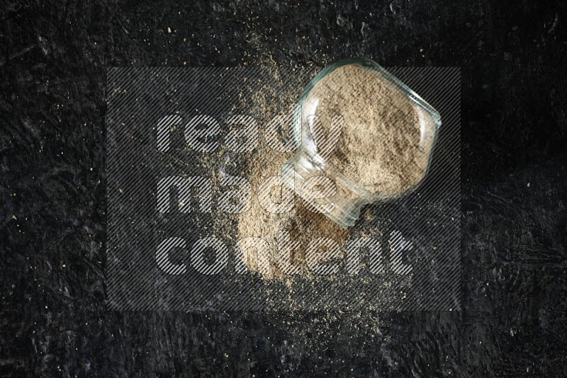 A flipped glass spice jar full of cardamom powder on textured black flooring