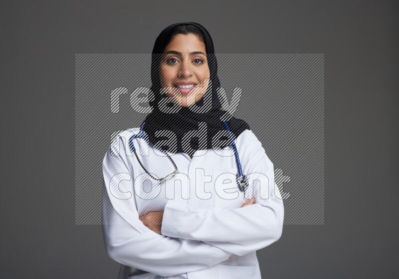 Saudi woman wearing lab coat with stethoscope standing with crossed arms on Gray background