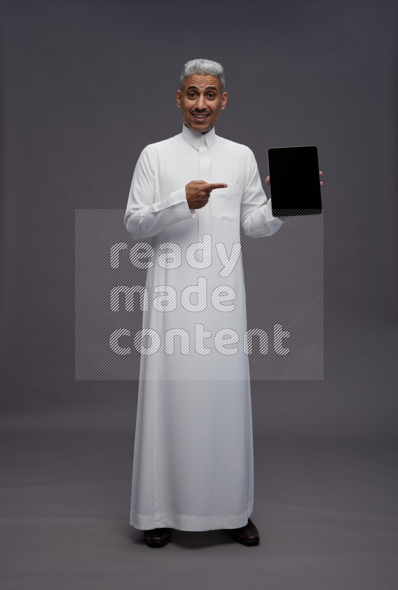 Saudi man wearing thob standing showing tablet to camera on gray background