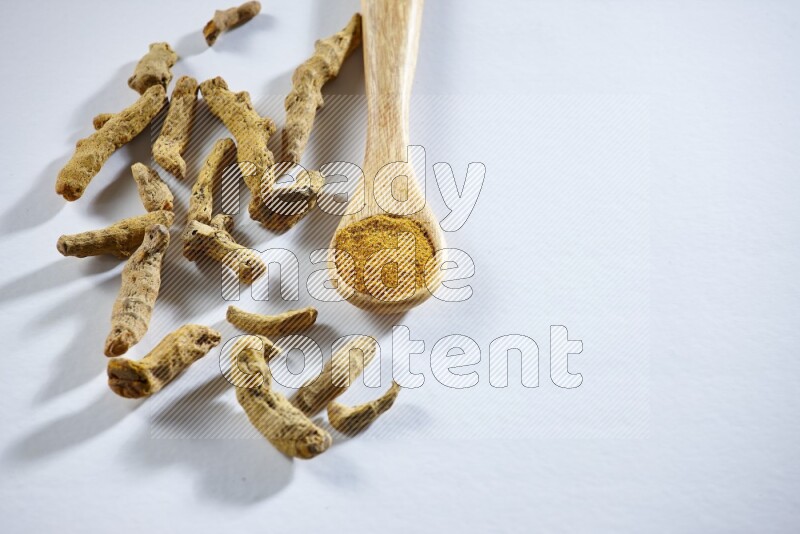 A wooden spoon full of turmeric powder with dried turmeric fingers beside it on white flooring