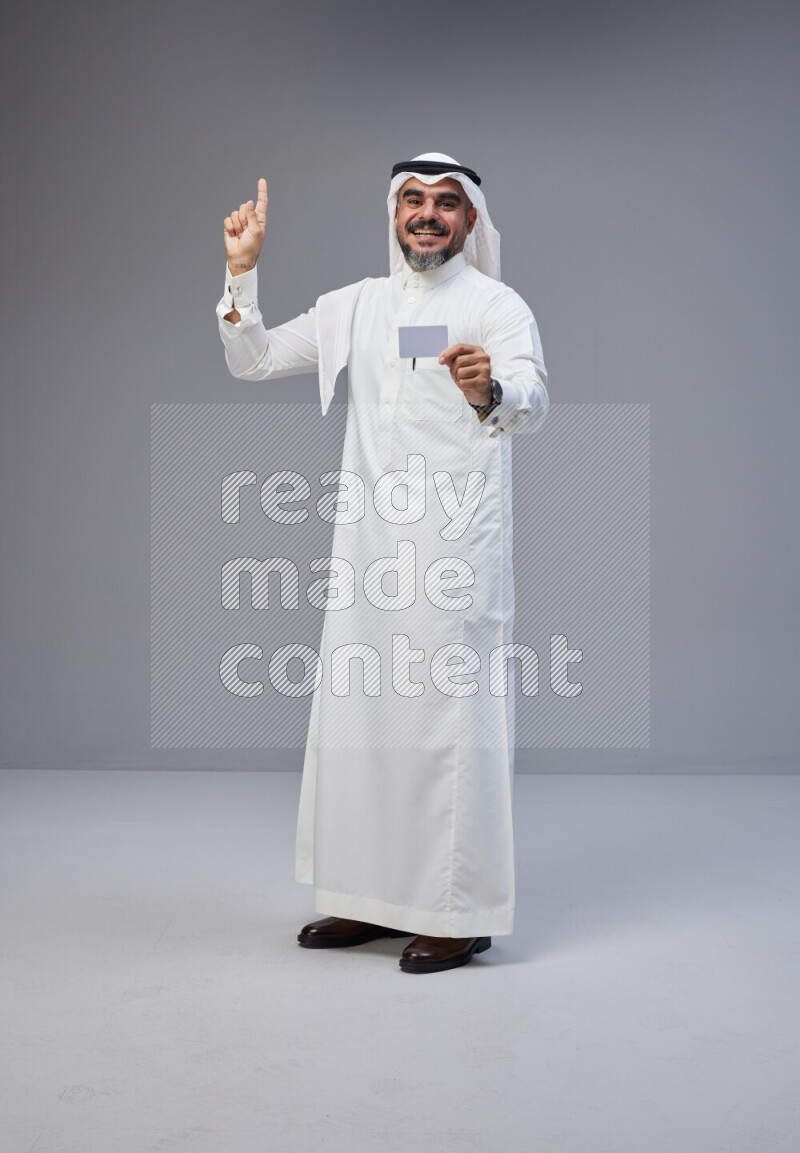 Saudi man Wearing Thob and white Shomag standing holding ATM card on Gray background