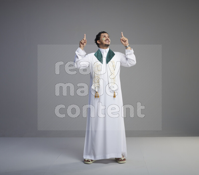 A Saudi man standing wearing thob and Saudi flag scarf with face painting on gray background