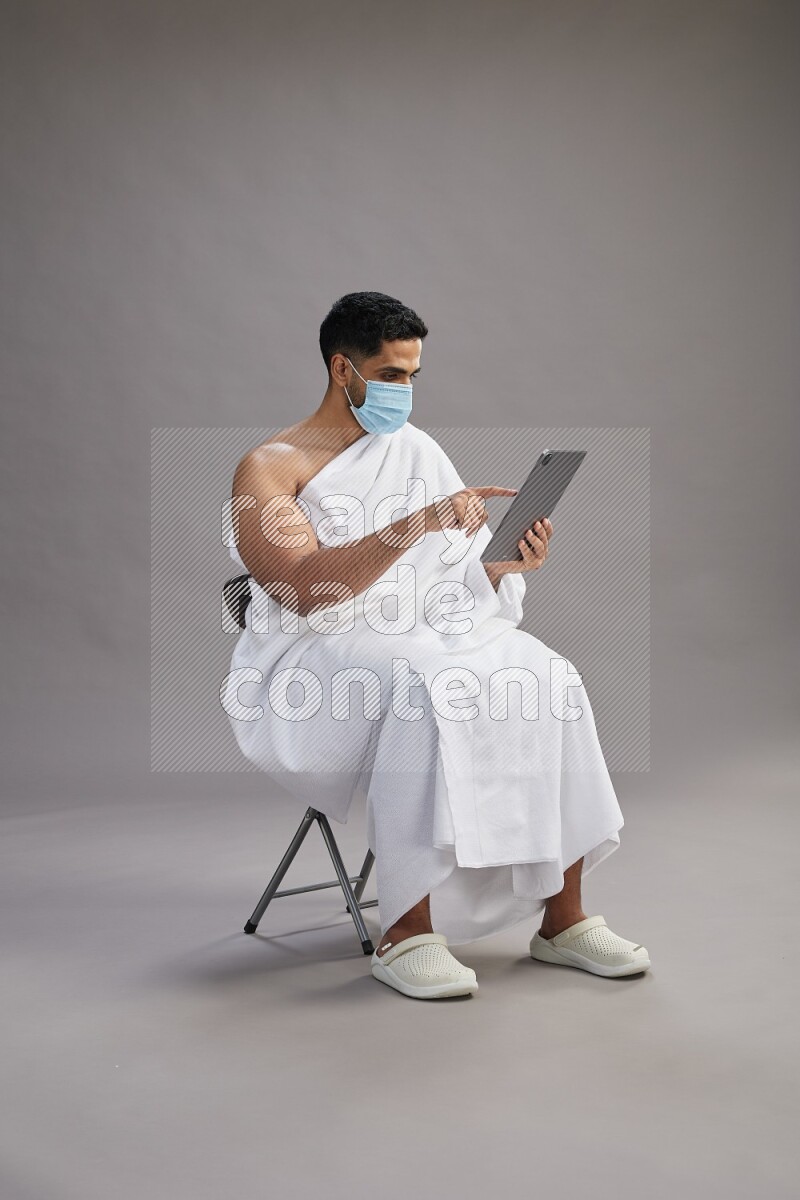 A man wearing Ehram with face mask sitting on chair holding a tablet on gray background