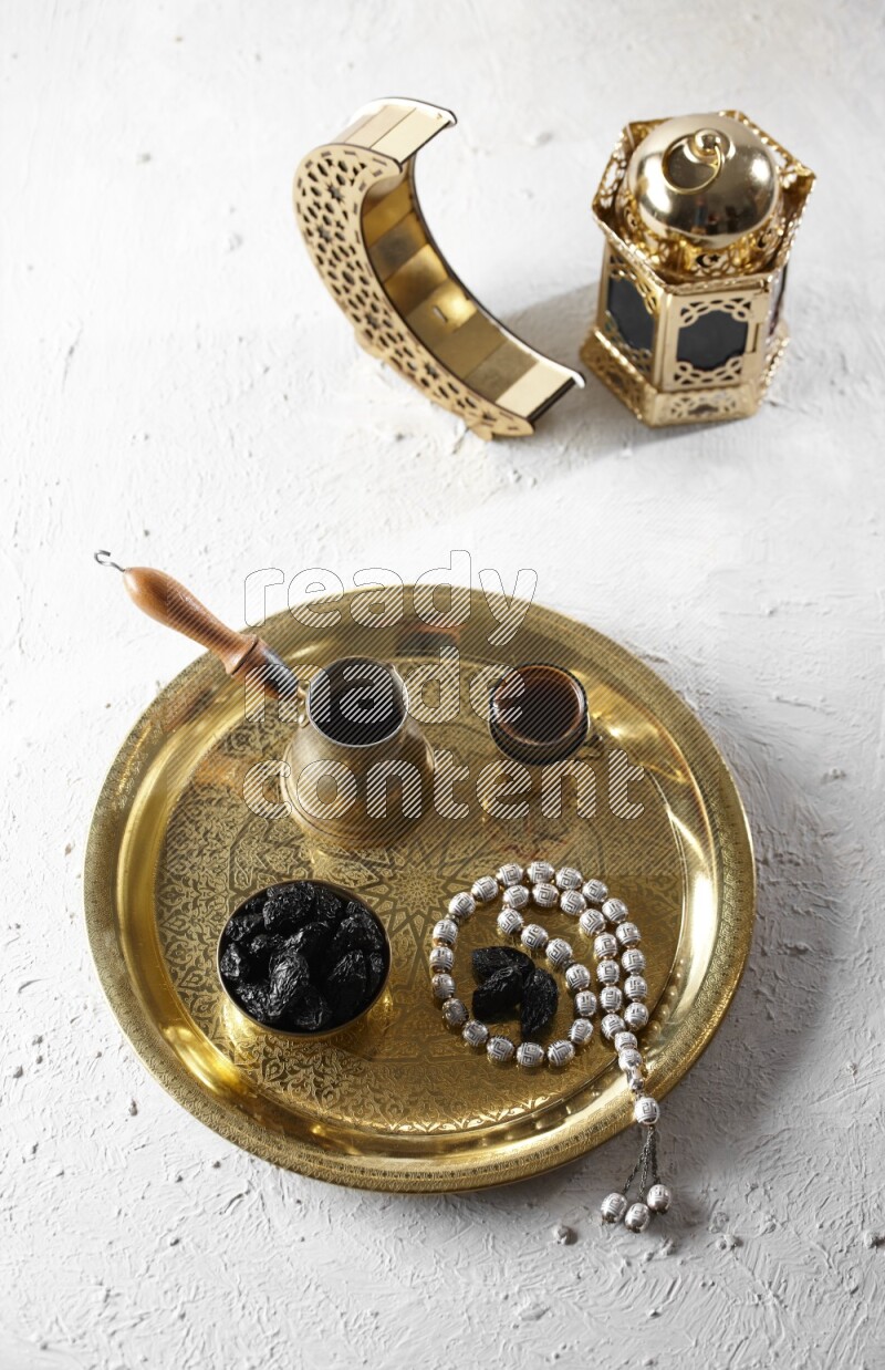 Dried plums in a metal bowl with coffee and prayer beads on a tray beside lanterns in a light setup