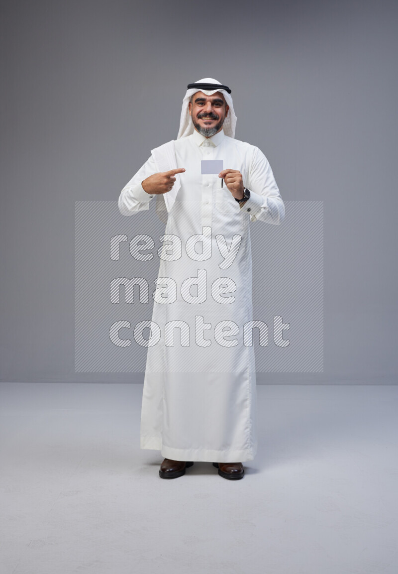 Saudi man Wearing Thob and white Shomag standing holding ATM card on Gray background