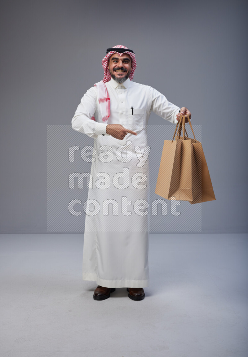 Saudi man Wearing Thob and red Shomag standing holding shopping bag on Gray background